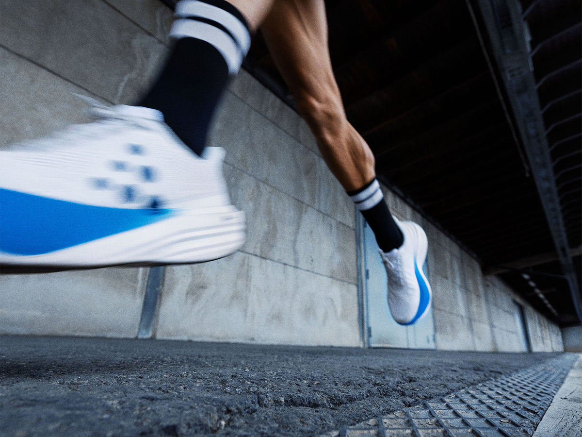 A close-up action shot of a runner's legs and feet wearing white running shoes with blue accents and black socks, captured on an urban street underpass.