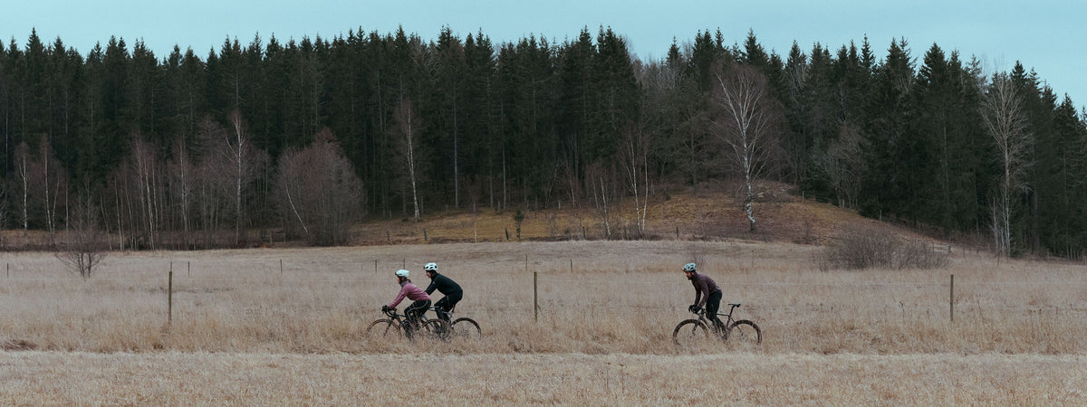 Three cyclists ride across a dry grass field with a forest in the background under a pale blue sky.