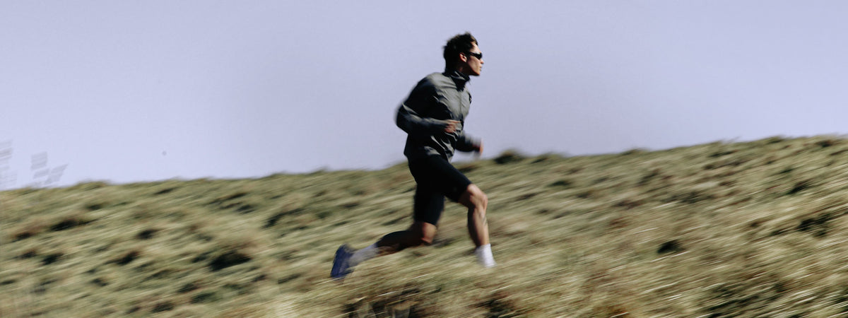A man running across a grassy hill wearing a dark gray long-sleeve athletic top, black shorts, white socks, and sunglasses under a pale sky.