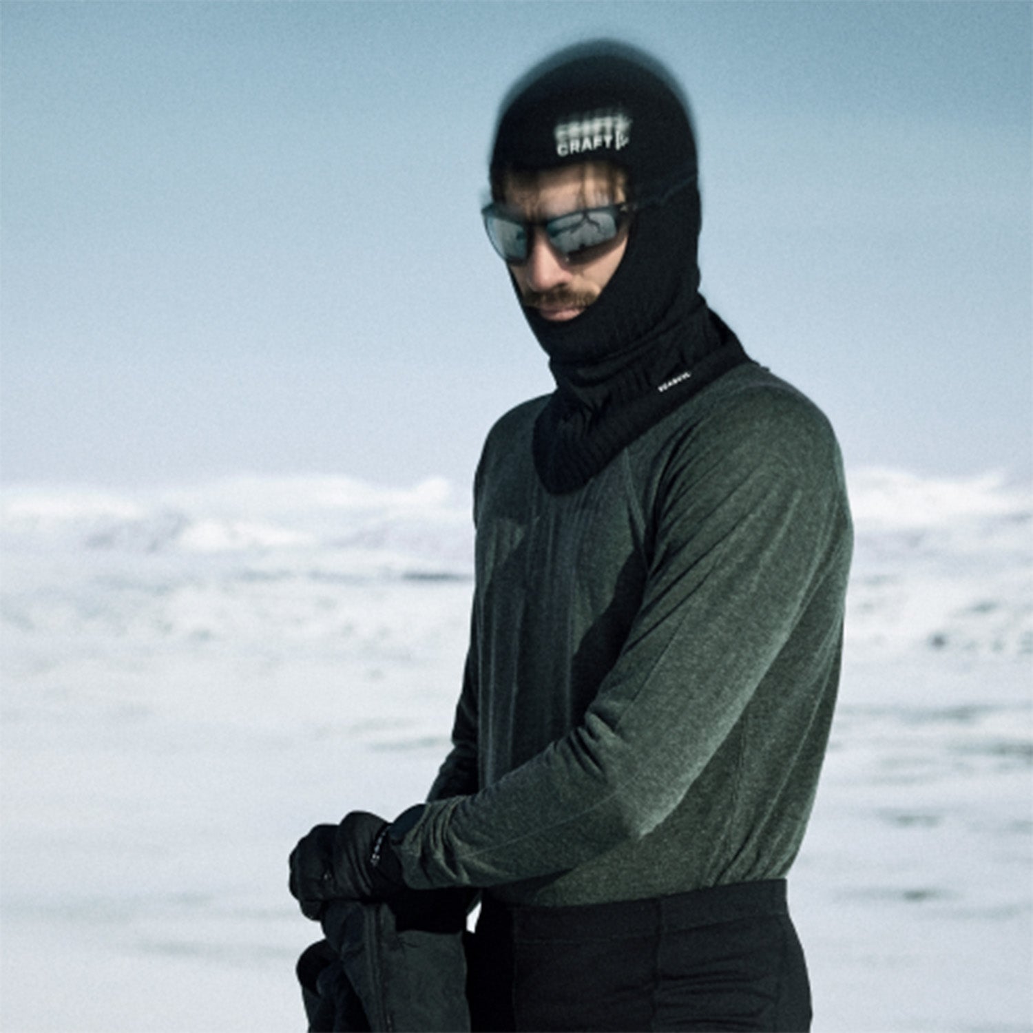 A man wearing a black ski balaclava, dark sunglasses, and a dark green long-sleeve technical shirt standing in a snowy outdoor environment.