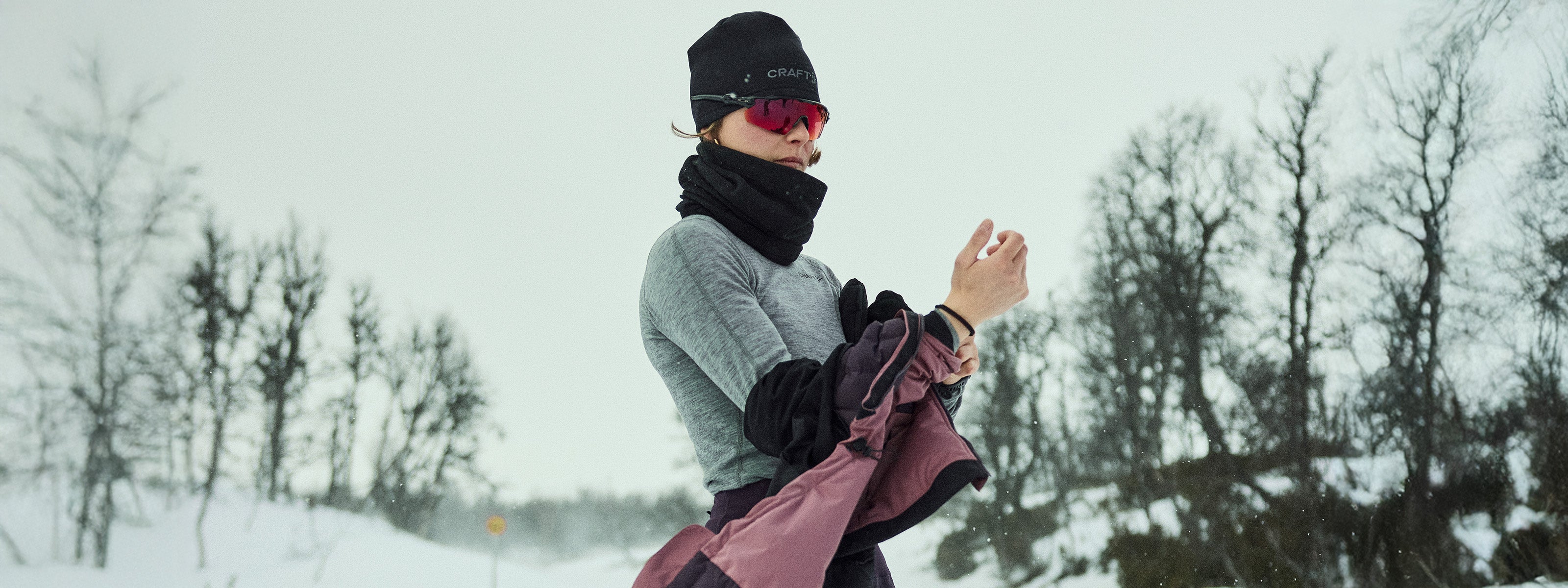 Person in winter clothing standing in a snowy landscape with bare trees.