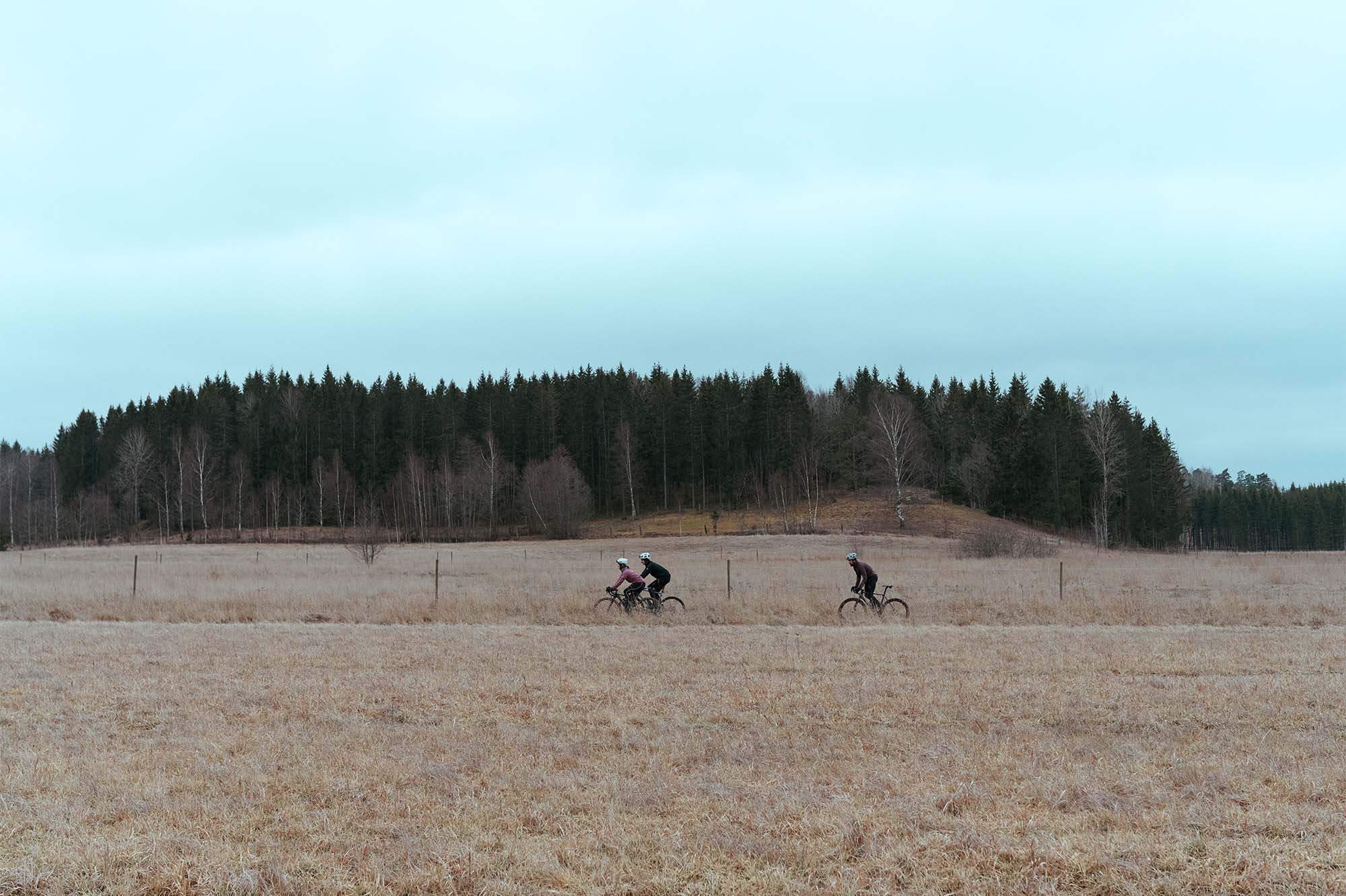 Two cyclists ride across a dry, grassy field under a cloudy sky, with a line of evergreen trees in the background.