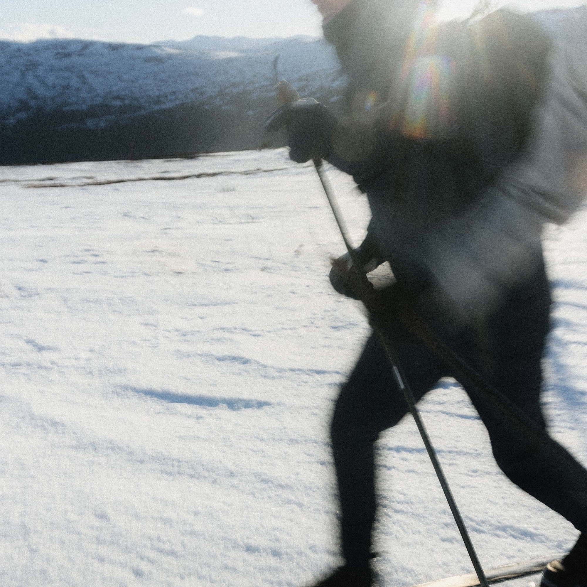 A skier in dark winter gear moves across a snowy mountain landscape with mountains and blue sky in the background. The photo is backlit by the sun, creating a bright flare and some motion blur.