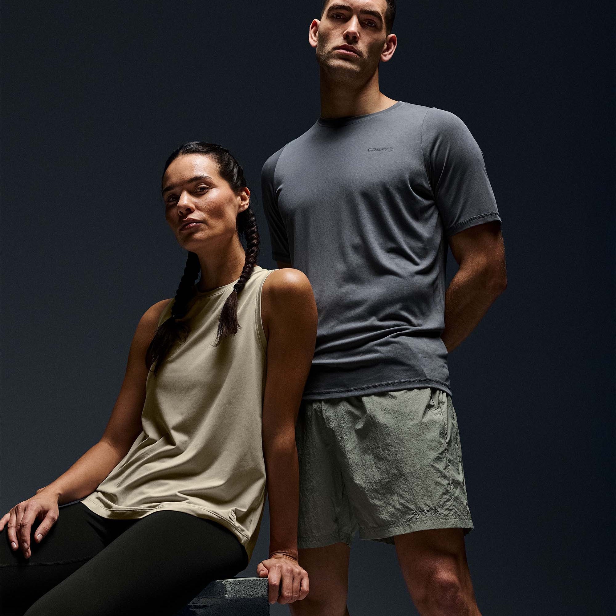 A man and a woman in athletic wear pose against a dark studio background. The man stands wearing a dark gray short-sleeve athletic t-shirt and light gray running shorts.
