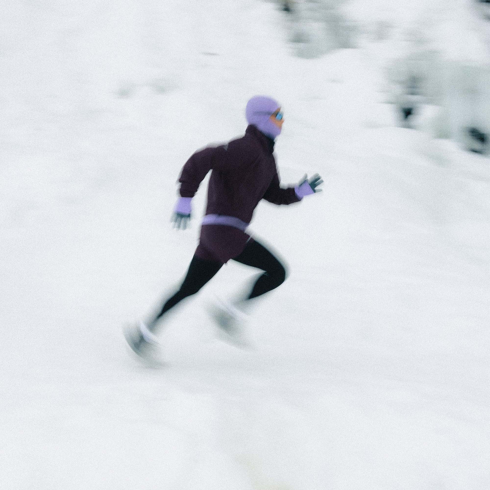 A person in a dark purple running outfit with a light purple helmet and gloves is captured in motion while skiing or snowboarding down a snowy slope.