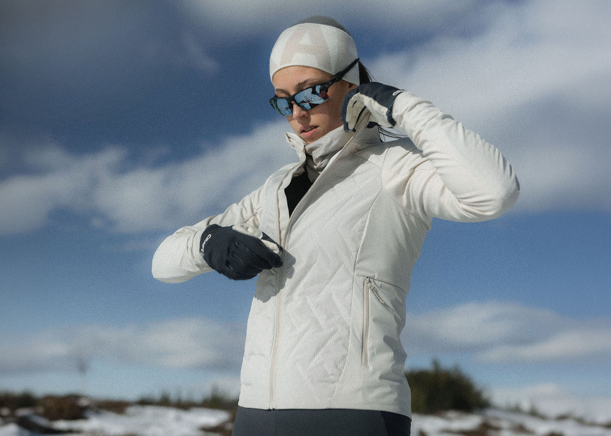 A woman wearing a white quilted winter jacket, matching white beanie, black gloves, and sunglasses stands outdoors against a blue sky with clouds and patches of snow.