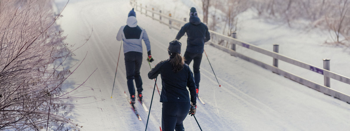 Three people cross-country skiing on a snowy path outdoors in winter clothing, with bare trees and a snowy landscape in the background.