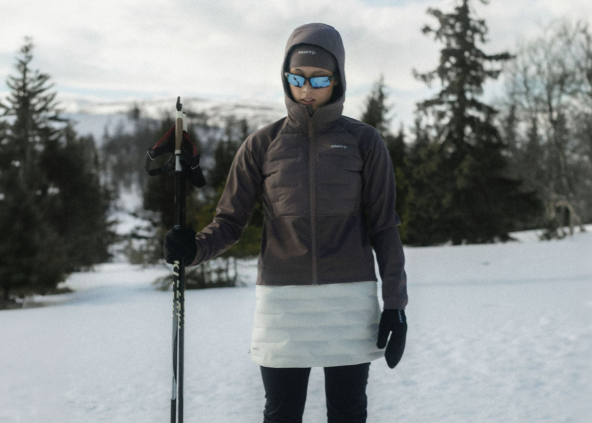 A person standing outdoors in the snow wearing a hooded insulated jacket in dark gray and white, with ski poles and sunglasses.