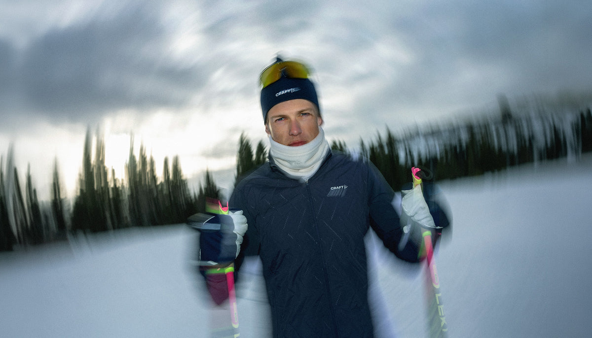 A skier wearing a dark navy technical jacket, a white neck gaiter, a black beanie with yellow goggles on top, and gloves, holding ski poles in a snowy outdoor setting with blurred trees and an overcast sky.