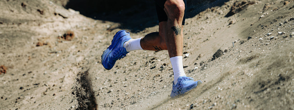 A close-up action shot of a runner's legs and feet wearing blue trail running shoes, white socks, and black shorts while running on a sandy, rocky trail.
