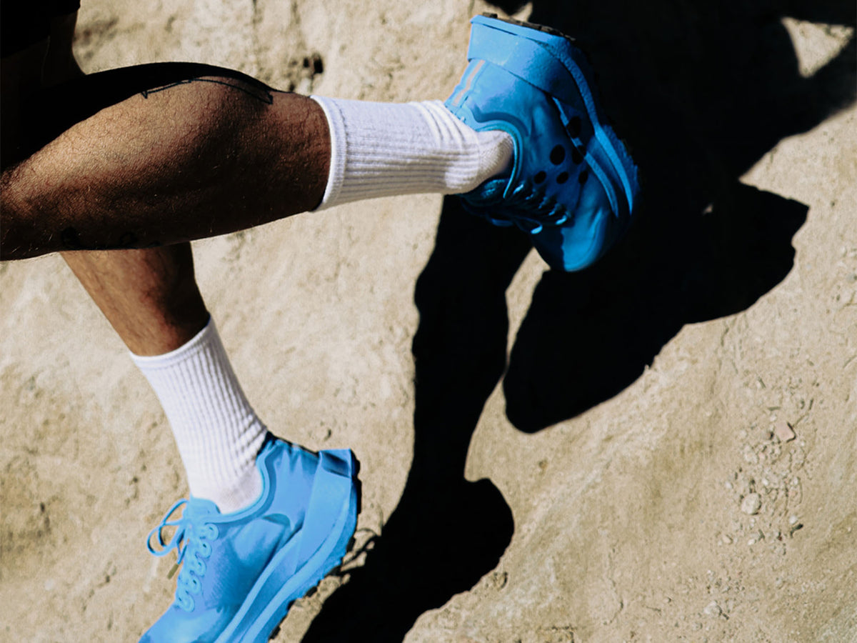 A close-up lifestyle shot of a person's legs and feet wearing bright blue athletic sneakers and white crew socks, standing on a sandy ground with strong shadows.