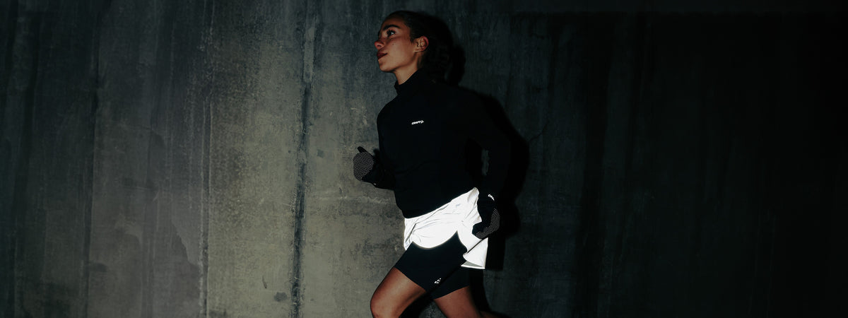 A woman running against a dark concrete wall, wearing a black long-sleeve athletic top and white shorts.