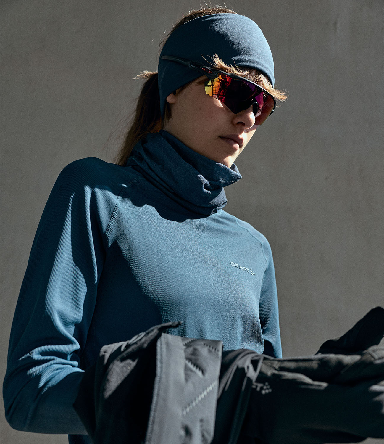 A woman wearing a slate blue athletic turtleneck top, matching headband, and mirrored sunglasses, holding a black jacket against a neutral gray background.