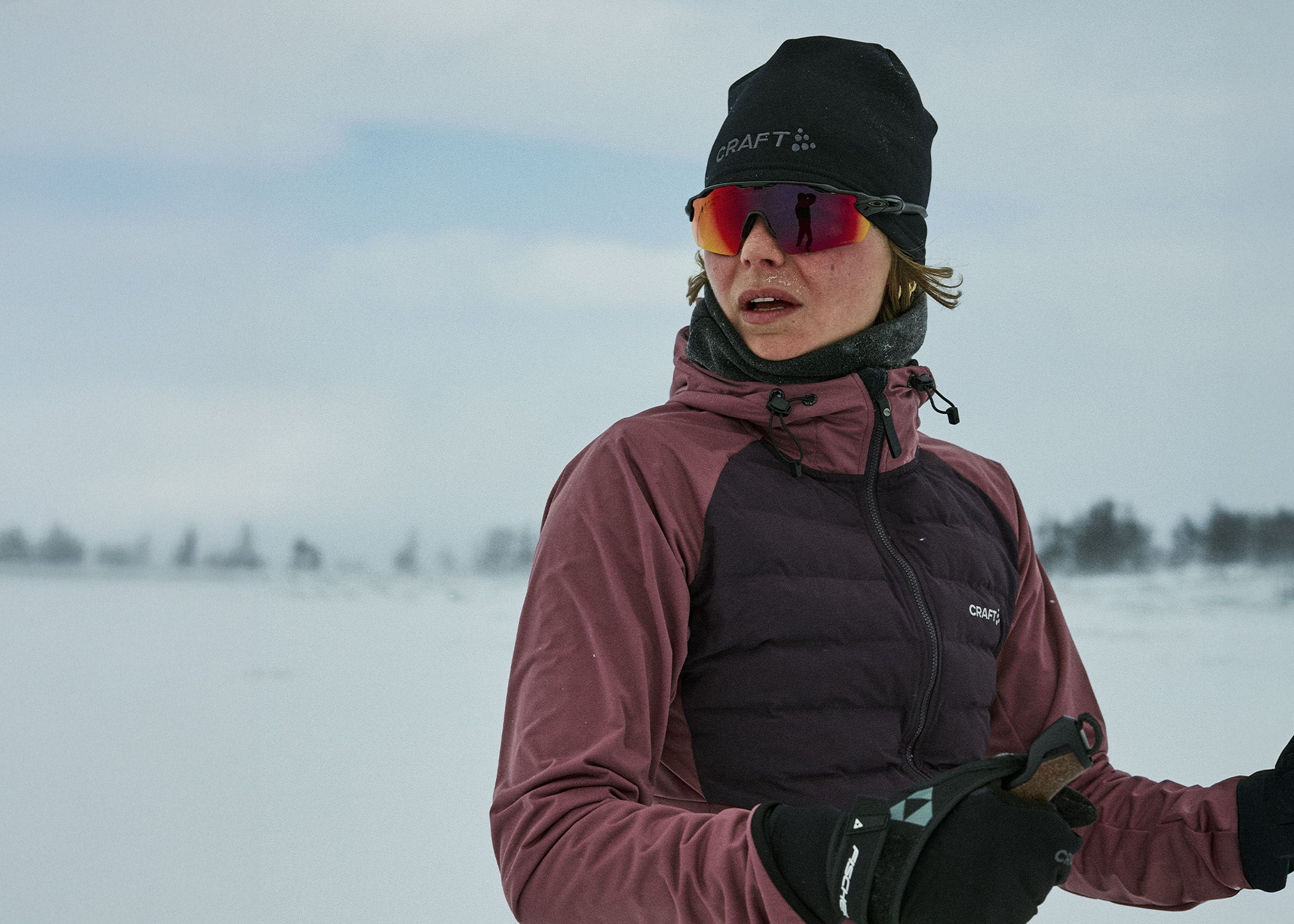 Personne portant une veste marron, un bonnet noir et des lunettes de soleil dans un paysage enneigé.