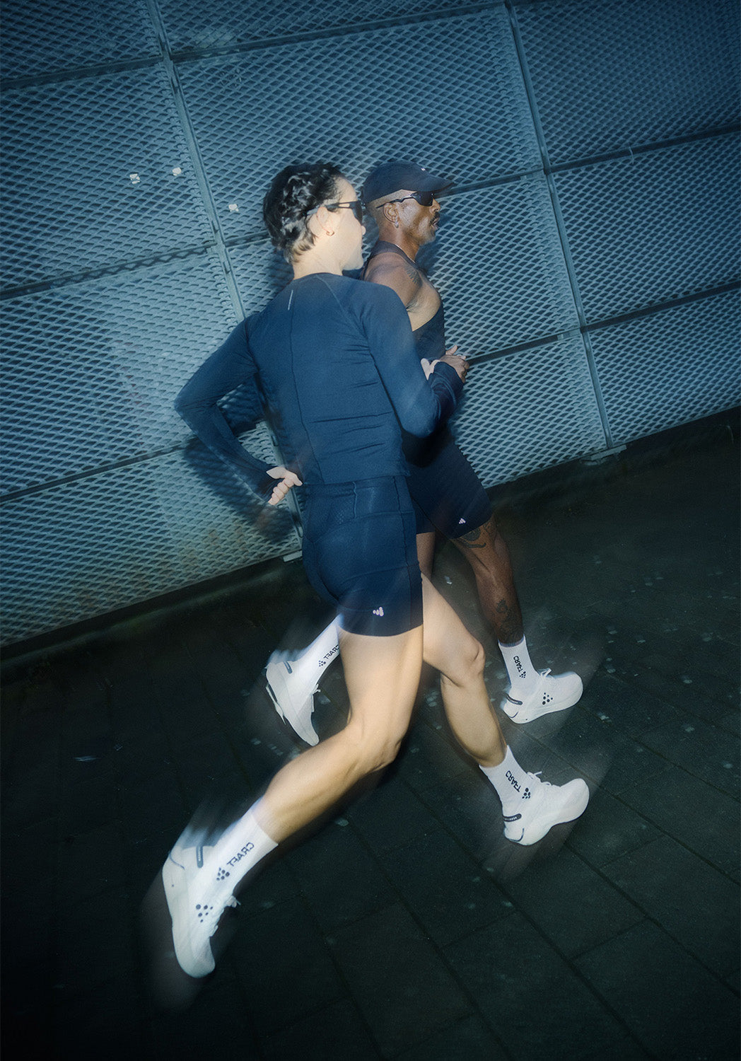 Deux personnes en tenue de sport courant sur une surface sombre avec un mur bleu texturé.