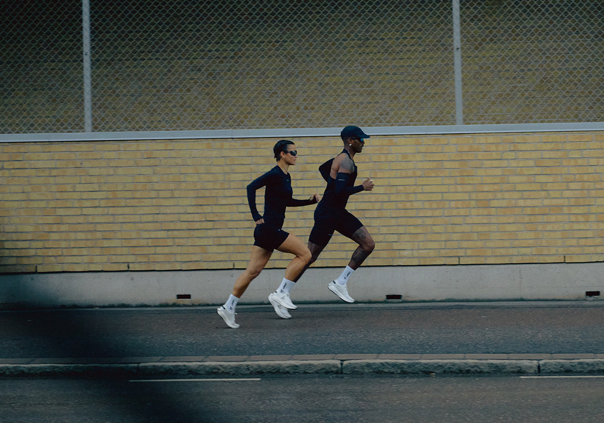 Two runners in athletic gear running on a street with a yellow brick wall in the background.