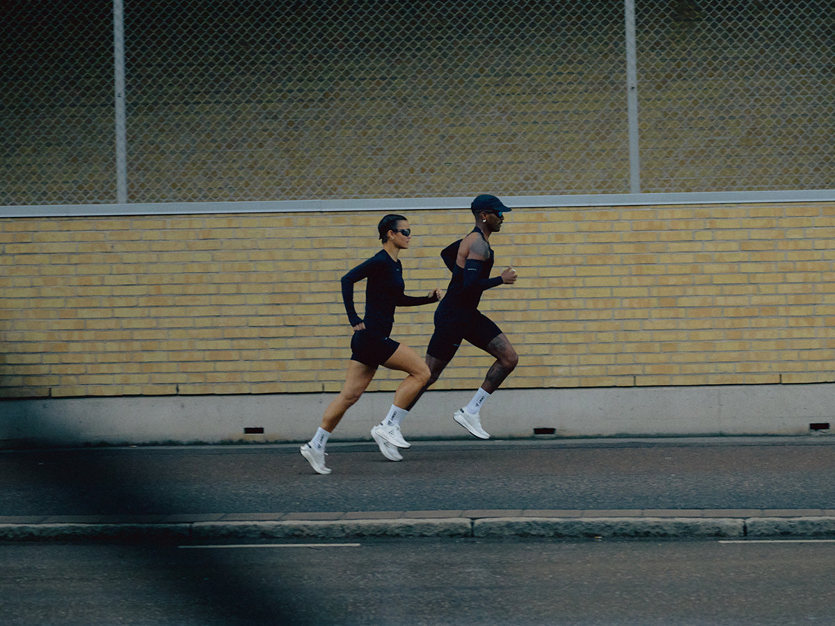 Two runners in athletic gear running on a street with a yellow brick wall in the background.