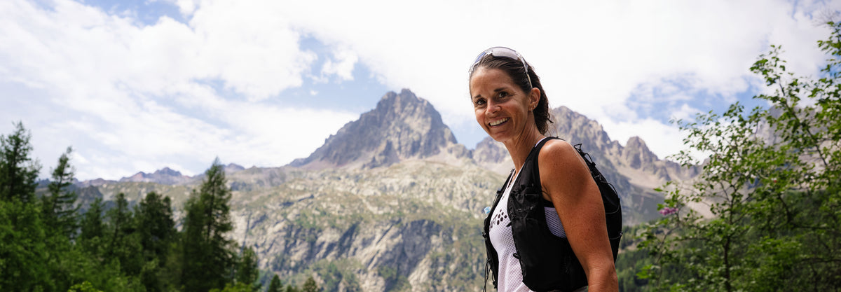 A woman wearing a black running vest over a white tank top stands outdoors in front of a mountain range under a cloudy sky, with trees visible in the foreground.