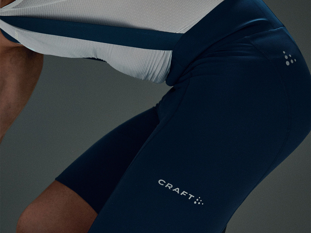 Close-up of a person wearing dark navy athletic cycling shorts with a white CRAFT logo and a white-and-navy jersey, cropped against a dark gray studio background.