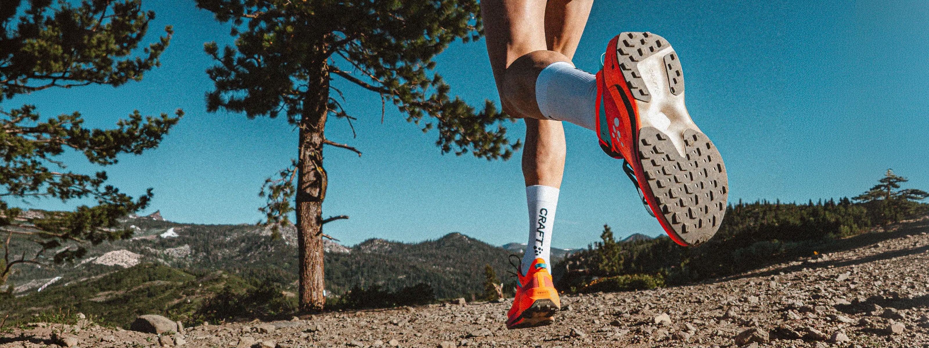 A close-up action shot of a runner's legs and feet mid-stride on an outdoor trail, showcasing a pair of bright orange and black running shoes, white socks with a black logo, and athletic shorts against a backdrop of pine trees and mountains under a clear blue sky.
