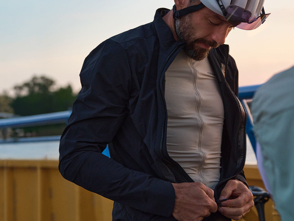 A man wearing a dark navy lightweight zip-up jacket over a beige cycling jersey, adjusting the zipper on his chest. He is wearing a cycling helmet with goggles, and the background shows a soft-focus outdoor setting at sunset.