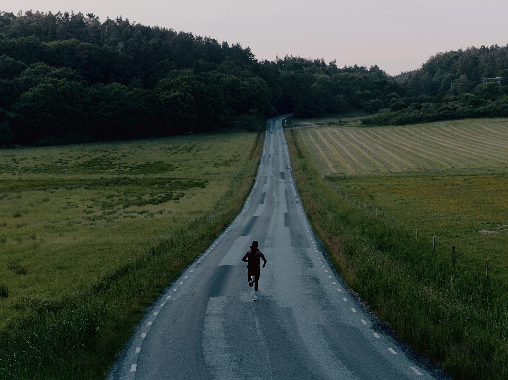 A lone runner runs down a narrow country road between green fields and a dark forest under a pale sky.
