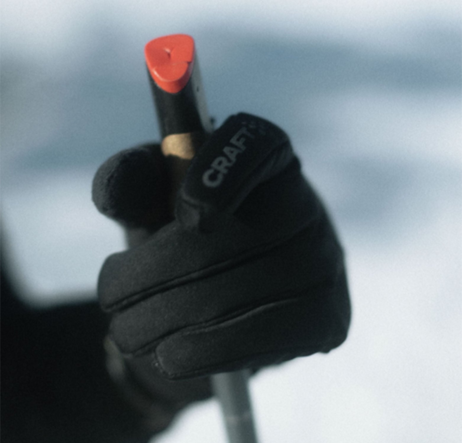 A close-up of a black Craft Glove holding a ski pole with a red and black tip against a blurred snowy background.