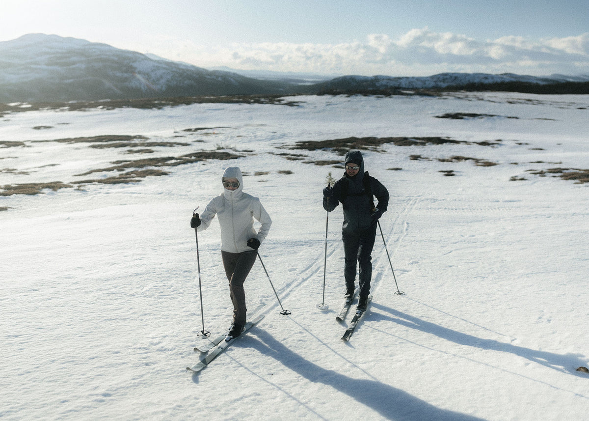Two people cross-country skiing on a snowy mountain landscape with hills and trees in the background.