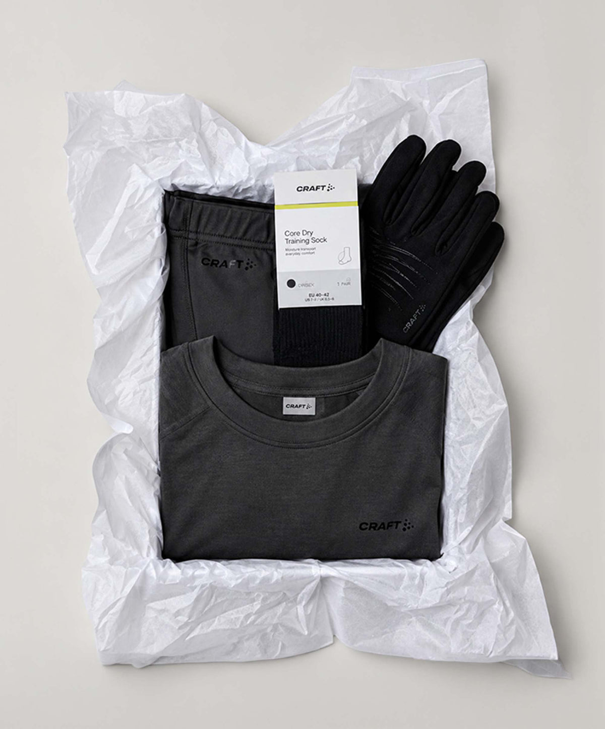 A flat-lay of a CRAFT brand multi-item gift set wrapped in crumpled white paper. The set includes a dark gray crewneck training shirt, black training shorts, black gloves, and a black training sock with its product tag still attached.