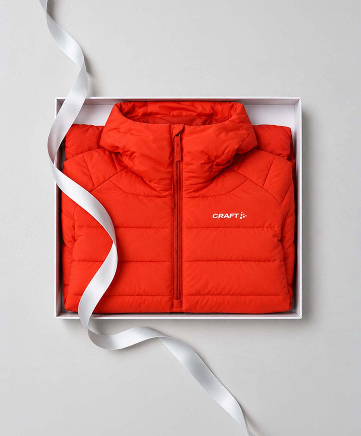 A bright red puffer jacket with a white CRAFT logo on the chest, neatly folded inside a white gift box with a white ribbon, set against a light gray background.