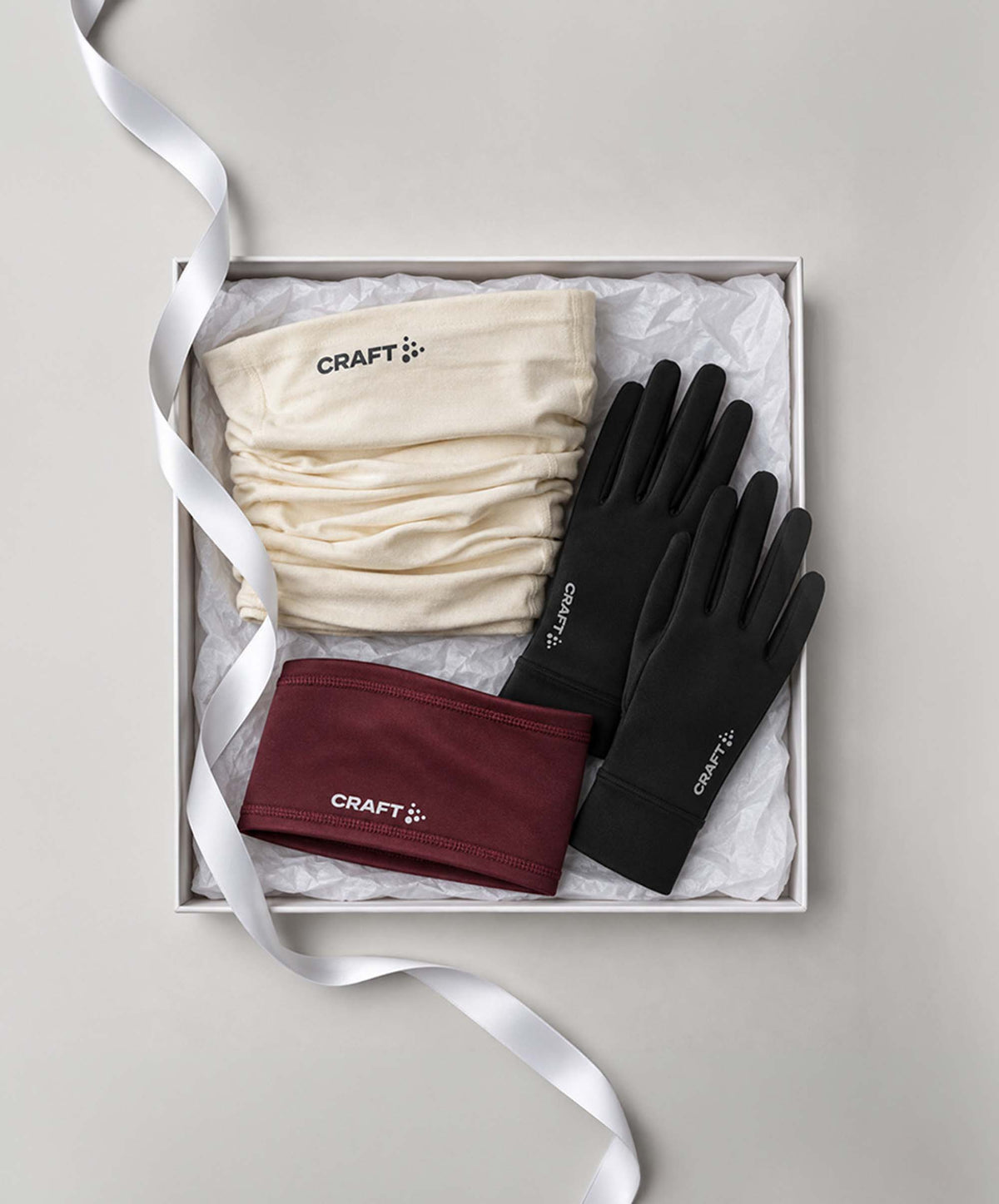 A gift set from Craft  in a white box with tissue paper and a white ribbon, containing a cream knit beanie, black touchscreen gloves, and a burgundy knit headband.