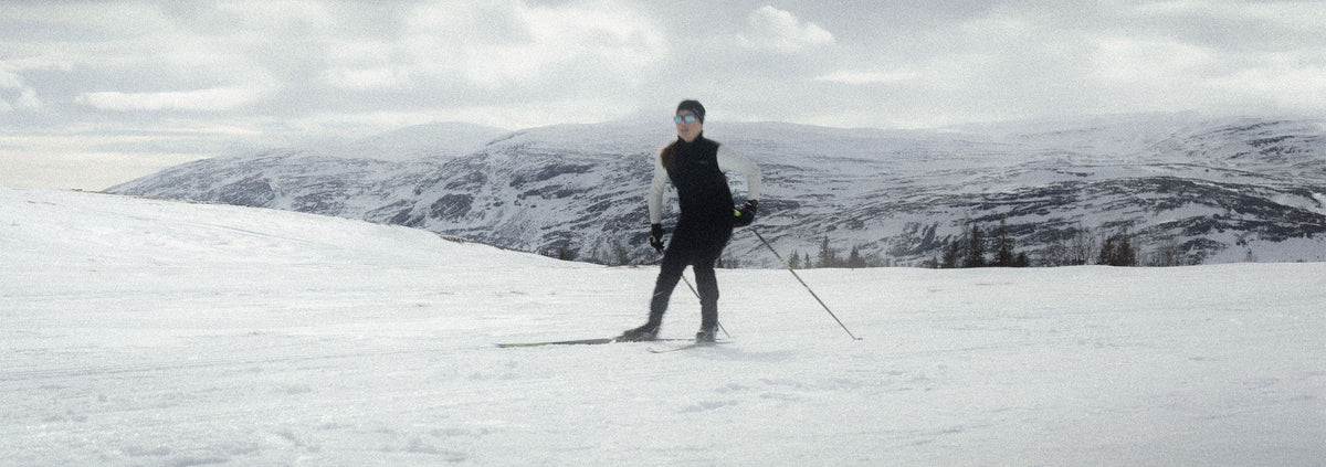 A person standing on skis in a snowy mountain landscape, wearing a black and white winter outfit with ski poles, under an overcast sky.
