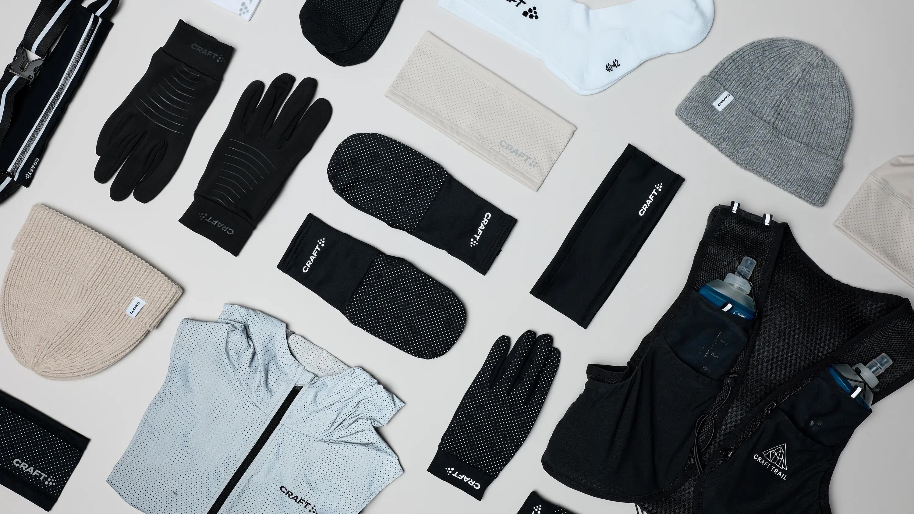A flat-lay arrangement of multiple outdoor and athletic accessories on a light gray background, including black gloves, a black neck gaiter, a black beanie, a light gray beanie, a light gray jacket, a black vest with a water bottle, and other matching gear.