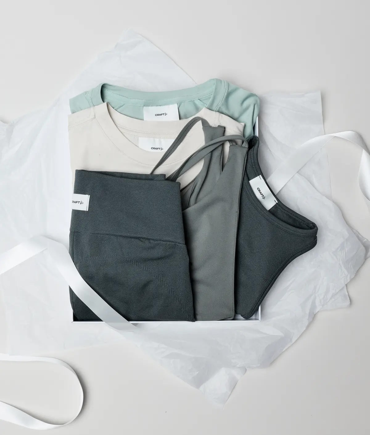 Three folded activewear sets in sage green, cream, and dark gray, arranged on white tissue paper with white ribbons. Each set includes a tank top and matching leggings.