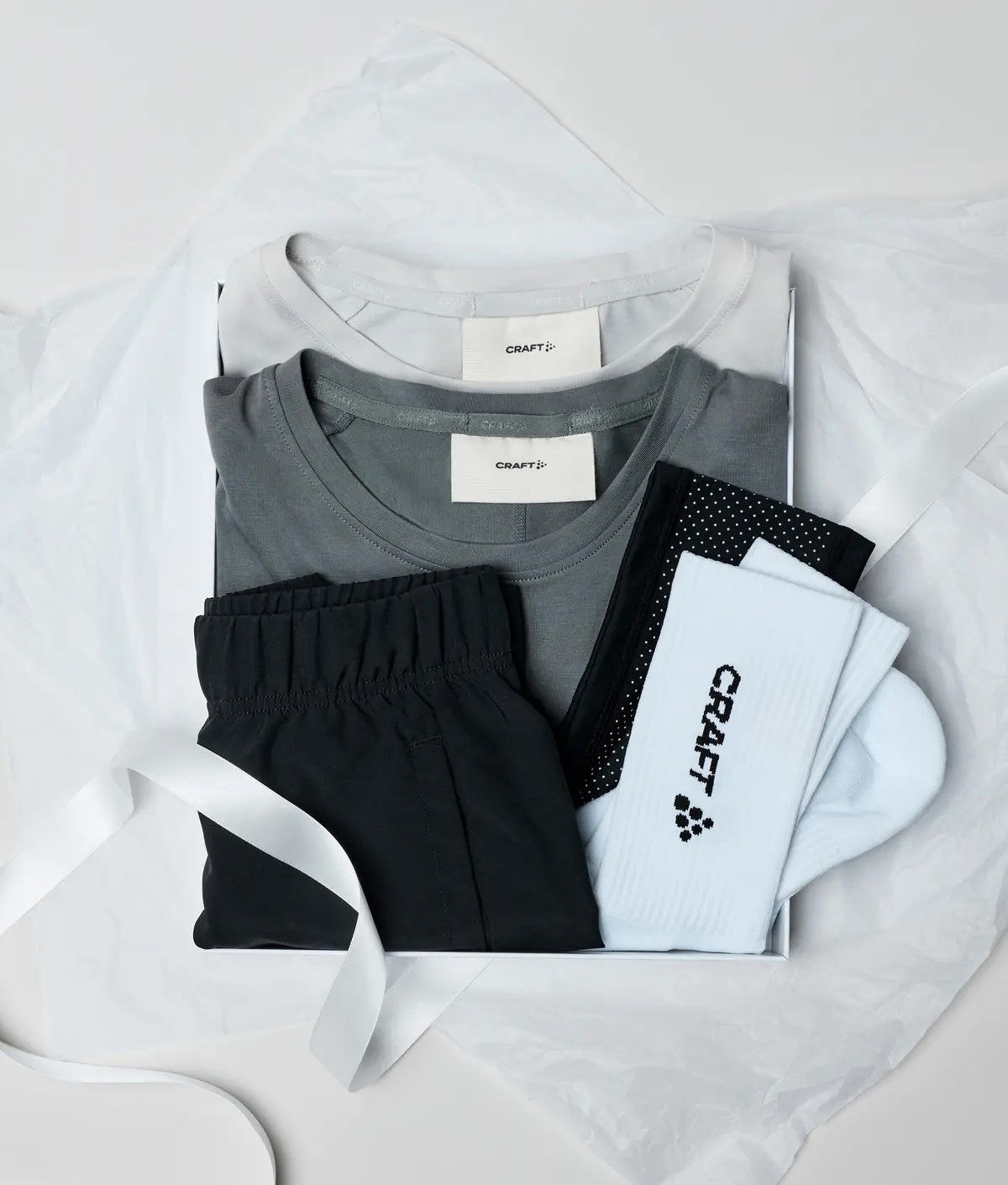 A flat-lay presentation of a CRAFT three-piece athletic set, including a gray short-sleeve t-shirt, black shorts, and light blue and black socks, arranged inside a white box with a white ribbon on a light gray background.